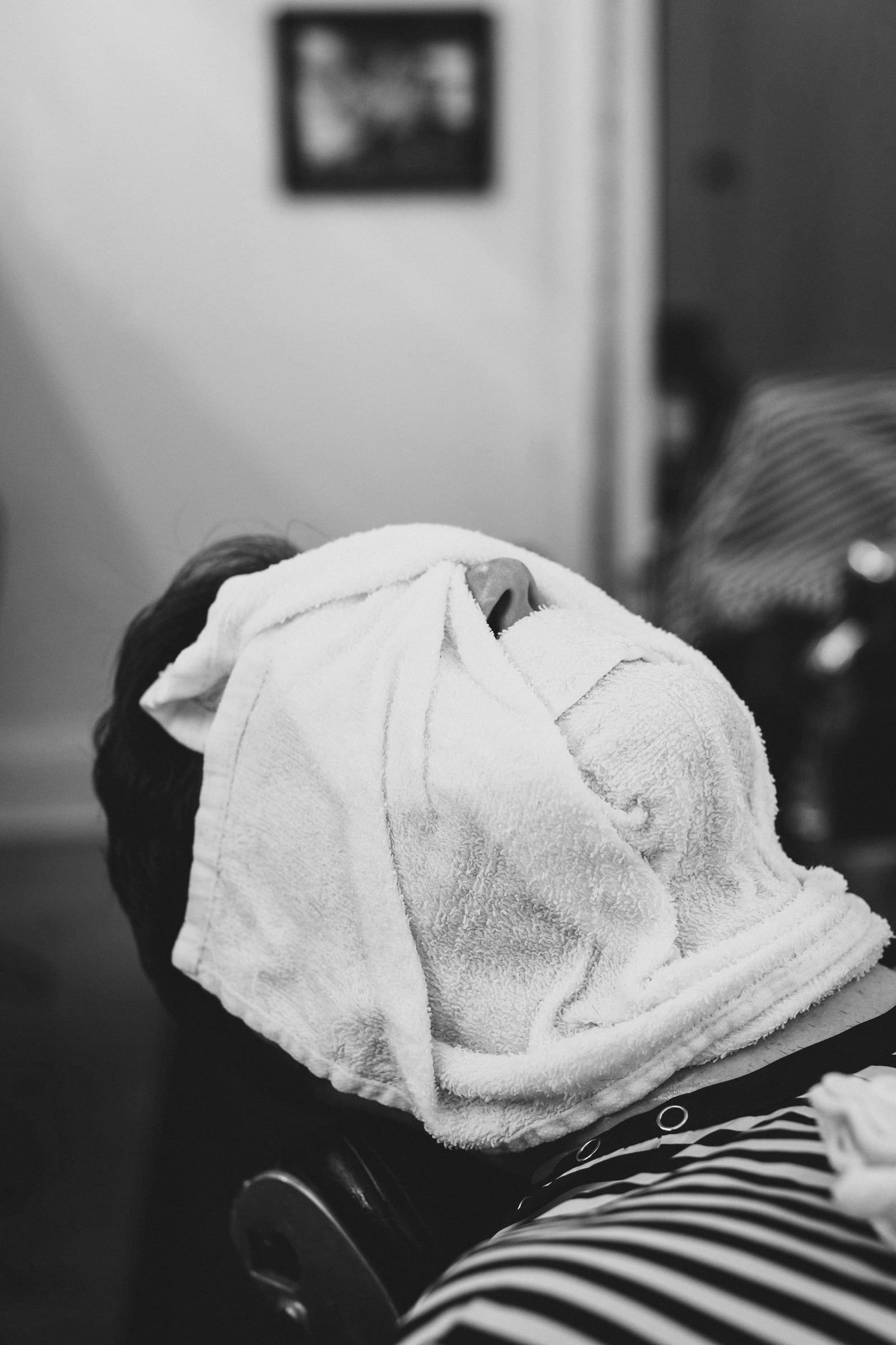 Gast liegt zurückgelehnt im Barberstuhl, das Gesicht unter einem heissen Tuch